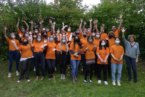Eine Gruppe von Studierenden stehen zusammen vor einer Baumgruppe. Sie tragen orangefarbene T-Shirts, Mund-Nasen-Bedeckungen und strecken ihre Arme in die Höhe