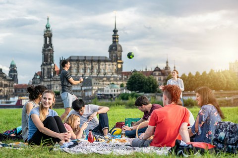 Foto: Gruppe von Studenten an der Elbe. Sie spielen Volleyball und sitzen plaudernd auf der Wiese.