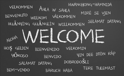 Die Darstellung zeigt eine schwarze Tafel. An diese wurde mit weißer Kreide groß das Wort "Welcome" in die Mitte geschrieben. Drum herum stehen viele kleinere Begriffe. Sie sind die Übersetzung dieses Wortes in verschiedenen Sprachen.