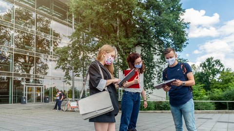 Das Foto zeigt drei Studierende auf dem Platz vor dem Hörsaalzentrum der TU Dresden. Alle drei Personen tragen einen Mund- und Nasenschutz. Sie Halten Notizbücher und Mappen in den Händen und reden miteinander.