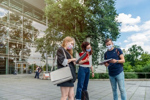 Das Foto zeigt drei Studierende auf dem Platz vor dem Hörsaalzentrum der TU Dresden. Alle drei Personen tragen einen Mund- und Nasenschutz. Sie Halten Notizbücher und Mappen in den Händen und reden miteinander.
