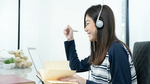 Das Foto zeigt eine junge Frau an ihrem Schreibtisch. Sie trägt Kopfhörer, lächelt und notiert etwas in ihrem Notizbuch. 