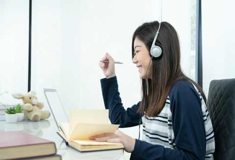 Das Foto zeigt eine junge Frau an ihrem Schreibtisch. Sie trägt Kopfhörer, lächelt und notiert etwas in ihrem Notizbuch. 