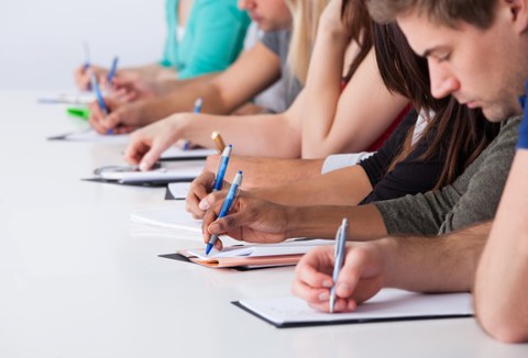 Studierende sitzen nebeneinander und schreiben konzentriert mit Stiften auf Papier.