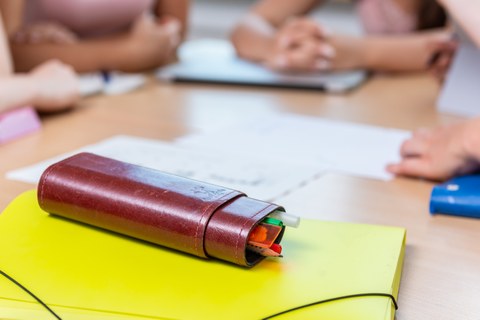 Ein rotbraunes Stiftmäppchen liegt auf einem gelben Hefter, im Hintergrund sind verschwommen junge Menschen an einem Tisch sitzend zu erkennen.