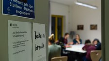 Ein Blick an der halb geöffneten Tür vorbei in den Raum der Schreibgruppe "Probieren geht über Studieren - Methoden ausprobieren": Die Teilnehmenden sitzen an Tischen und sprechen miteinander.