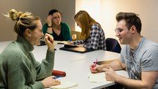 Vier Studierende sitzen an einem Tisch. Sie haben jeweils einen Block und einen Stift vor sich und sprechen in Zweiergruppen miteinander.