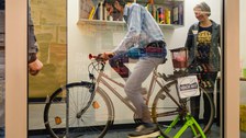 Hinter einer Glasscheibe steht ein aufgeFahrrad, auf dessen Gepäckträger ein Mixer montiert ist. Ein Mann sitzt auf dem Rad