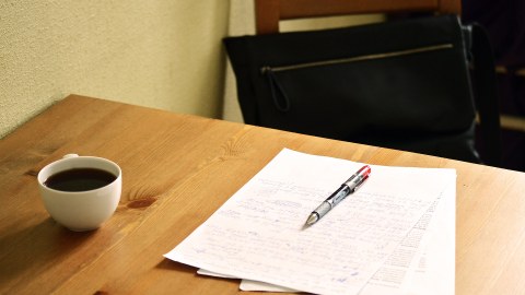 Das Foto zeigt die Ecke eines Holztisches, vor welchem ein leerer Stuhl steht. Auf dem Tisch liegen drei beschriebene Blatt Papier aufeinander, darauf liegt ein Kugelschreiber. Links daneben steht eine Tasse mit Kaffee. Die Sonne scheint auf den Arbeitsplatz.