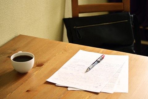 Das Foto zeigt die Ecke eines Holztisches, vor welchem ein leerer Stuhl steht. Auf dem Tisch liegen drei beschriebene Blatt Papier aufeinander, darauf liegt ein Kugelschreiber. Links daneben steht eine Tasse mit Kaffee. Die Sonne scheint auf den Arbeitsplatz.