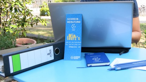 On a table is an opened laptop, in front of it leans a bookmark with "Schreibberatung! Is it working for you?", to the right of it are pencils, to the left a folder.