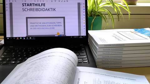 A laptop with the open PDF of the writing didactics starter guide, in front of it the open print copy and next to it a stack of closed print copies.