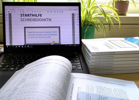 A laptop with the open PDF of the writing didactics starter guide, in front of it the open print copy and next to it a stack of closed print copies.