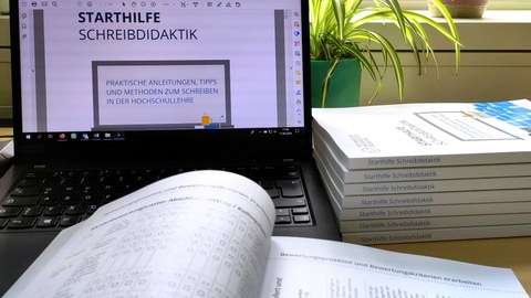 A laptop with the open PDF of the writing didactics starter guide, in front of it the open print copy and next to it a stack of closed print copies.