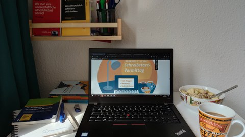 A laptop stands open on a desk, with a graphic on it that reads "2.09. 7-12 a.m.: Shrine Start Morning." In the background, books for scientific reading and writing.