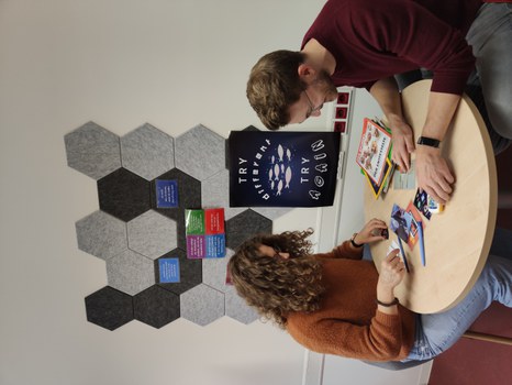 Two people are sitting at a round table, looking at newspapers and puzzles on the table. In the background: a pinboard with postcards and a poster with the text: “Try different. Try again.”