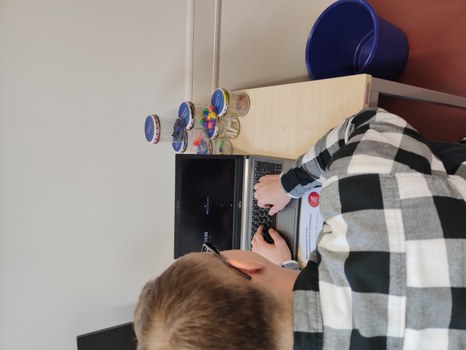 A table with a laptop with a black screen, in front of it a man in a checked shirt typing on the keyboard. Behind the laptop: screw-top jars stacked in a pyramid with various contents: paper clips, rolls of paper, pins, ink cartridges, interdental cleaners.