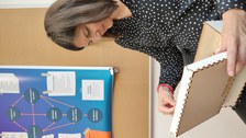A woman sits on a chair with an open wooden box in her hand. In the box: puzzle-like wooden pieces. Behind it: Pinboard with a poster of a flow chart, contents not recognizable.