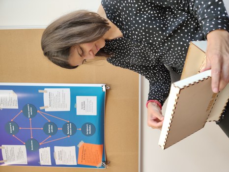 A woman sits on a chair with an open wooden box in her hand. In the box: puzzle-like wooden pieces. Behind it: Pinboard with a poster of a flow chart, contents not recognizable.