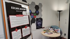 Part of a room, in the foreground: “Escape Room - Mika's Dream” poster with “Your Mission” information boxes and clues that cannot be read. Behind it: postcards on a pinboard, a table with newspapers, pens and puzzles, an armchair with cushions.