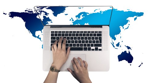 Draufsicht eines Laptops mit Händen auf der Tastatur. Der Desktop zeigt den Ausschnitt einer eurozentristischen Weltkarte. Dieser Ausschnitt wird im Hintergrund, um den Laptop herum, weitergeführt.