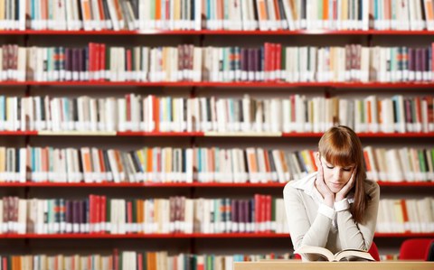 Foto einer jungen Studentin, die vor einer Bücherwand an einem Tisch sitzt und ein Buch liest.