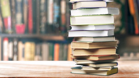 The photo shows a table with a large stack of books. In the background a bookcase can be seen blurred.