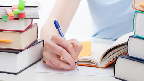 The photo shows the upper body of a young person writing on a sheet of paper. To the left and right of the paper: stacks of books with many colorful notes.