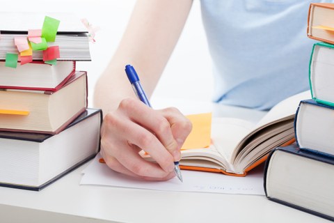 The photo shows the upper body of a young person writing on a sheet of paper. To the left and right of the paper: stacks of books with many colorful notes.
