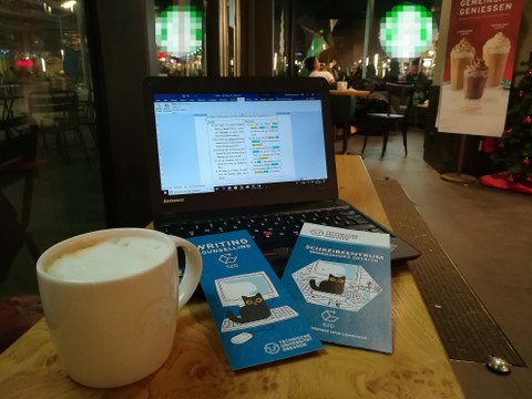 Ein Arbeitsplatz in einem bekannten Coffeeshop, zu sehen ist ein geöffneter Laptop, darauf Flyer des Schreibzentrums, links eine Tasse Kaffee, im Hintergrund Cafébesucher.