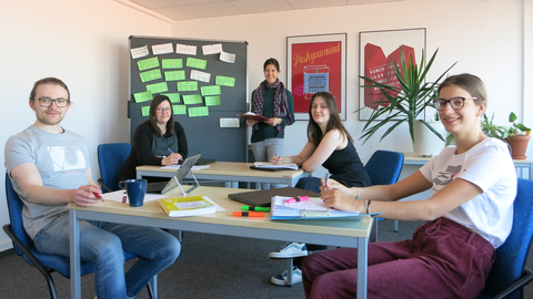 The four newly trained writing tutors sit across from each other at tables with laptops and smile into the camera. In the background is Nina Melching, head of the Writing Center.