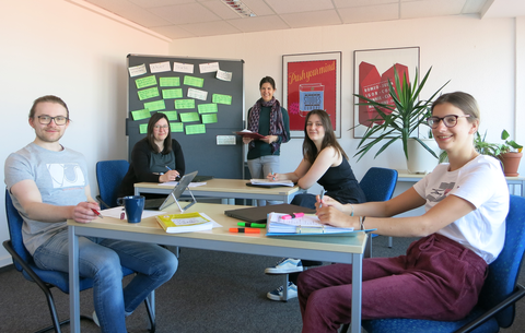 The four newly trained writing tutors sit across from each other at tables with laptops and smile into the camera. In the background is Nina Melching, head of the Writing Center.