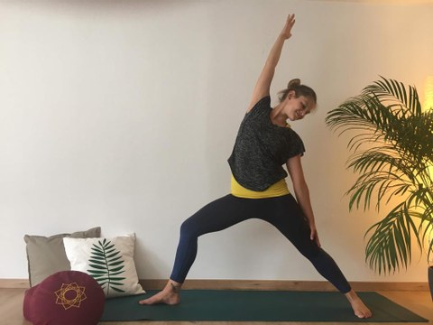 The photo shows a young woman standing on a yoga mat with her legs up, her right arm pointing upwards, the left resting on her thigh. In the background is a palm tree, cushions lie on the floor.