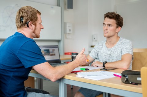 Das Foto zeigt zwei Studenten in einem Seminarraum. Sie sitzen sich gegenüber und sprechen miteinander. Zwischen ihnen liegen Zettel und Stifte. Einer der beiden Studenten trägt ein Hörgerät. 