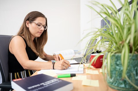  Das Foto zeigt eine Studentin an ihrem Schreibtisch. Vor ihr steht ihr Laptop und viele weitere Büromaterialien. Mit einem Stift notiert sie gerade etwas auf einem Block.