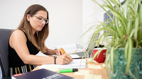  Das Foto zeigt eine Studentin an ihrem Schreibtisch. Vor ihr steht ihr Laptop und viele weitere Büromaterialien. Mit einem Stift notiert sie gerade etwas auf einem Block.
