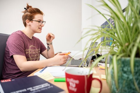 Das Foto zeigt eine Studentin an ihrem Schreibtisch. Vor ihr steht ihr Laptop und viele weitere Büromaterialien. Sie hält einen Stift in der Hand und schaut auf den Laptop. 