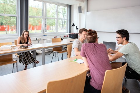 Das Foto zeigt mehrere Studierende in einem Seminarraum. Im Vordergrund sitzen sich drei Studierende gegenüber und unterhalten sich. Im Hintergrund sitzt eine Studentin und schaut auf ihr Smartphone. 