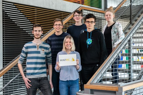 Six first-year students, two women and four men, stand on a staircase. One woman in the foreground holds a sign on which MINT-EC is written, all six receive a scholarship at the TU Dresden. 