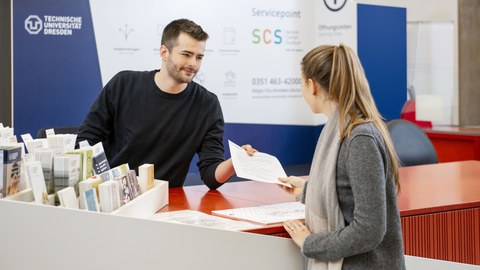 At the service point of the Service Center Studium, an employee hands a flyer to a student. The employee smiles. On the left side of the picture you can see a display with different flyers.