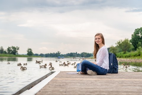 Eine Studentin sitzt auf einem Steg an der Elbe. Vor ihr liegen Bücher. Im Hintergrund sind Enten zu sehen. 