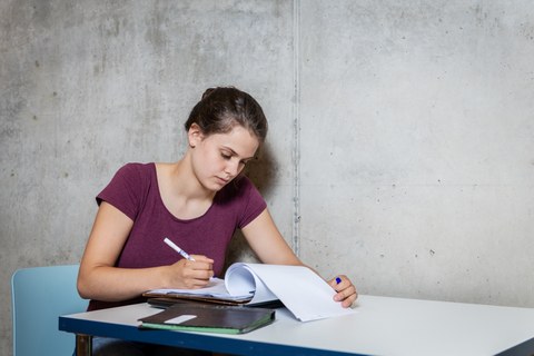 A female student is sitting at a table on a chair in the library. In front of her there are books. She is writing with a pen in a notebook. Her facial expression is concentrated. 