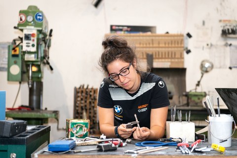 A student is sitting at a workbench. On the workbench and in the background you can see many different tools. The student is looking concentrated on the tool she is holding in her hand. 