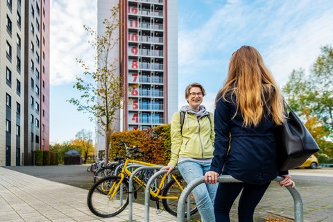 The photo shows two female students leaning against bicycle stands and talking. In the background are bicycles and two large student dormitories. 