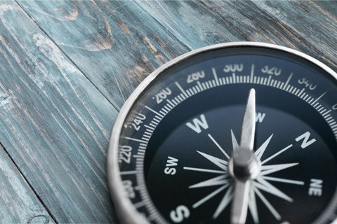 Das Foto zeigt einen Kompass, der sich auf einer Holzunterlage befindet. 