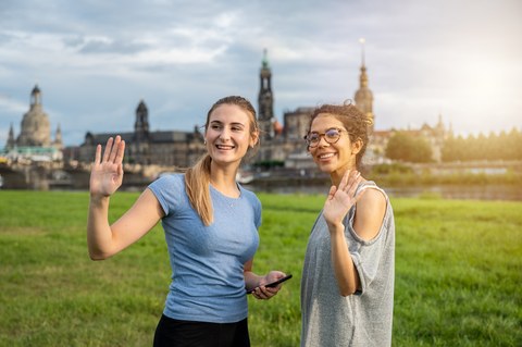 Zwei Studentinnen stehen an der Elbwiese in Dresden und winken.