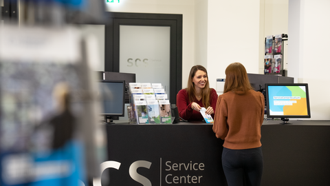 Foto des Empfangsbereich des ServiceCenterStudium: zwei Frauen stehen an der schwarzen Theke, eine davor uns abgewandt, die andere dahinter lächelnd und einen Flyer in der Hand haltend, erklärend.