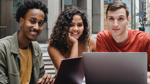 Drei Studierende sitzen vor einem Laptop. Links ist ein Mann, in der Mitte eine junge Frau und rechts ebenfalls ein junger Mann.