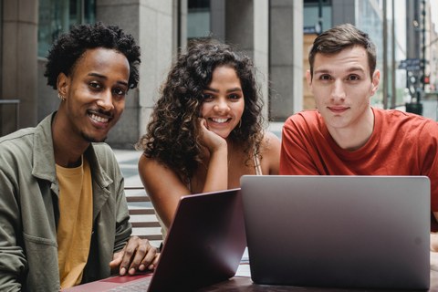 Drei Studierende sitzen vor einem Laptop. Links ist ein Mann, in der Mitte eine junge Frau und rechts ebenfalls ein junger Mann.