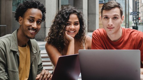 Drei Studierende sitzen vor einem Laptop. Links ist ein Mann, in der Mitte eine junge Frau und rechts ebenfalls ein junger Mann.